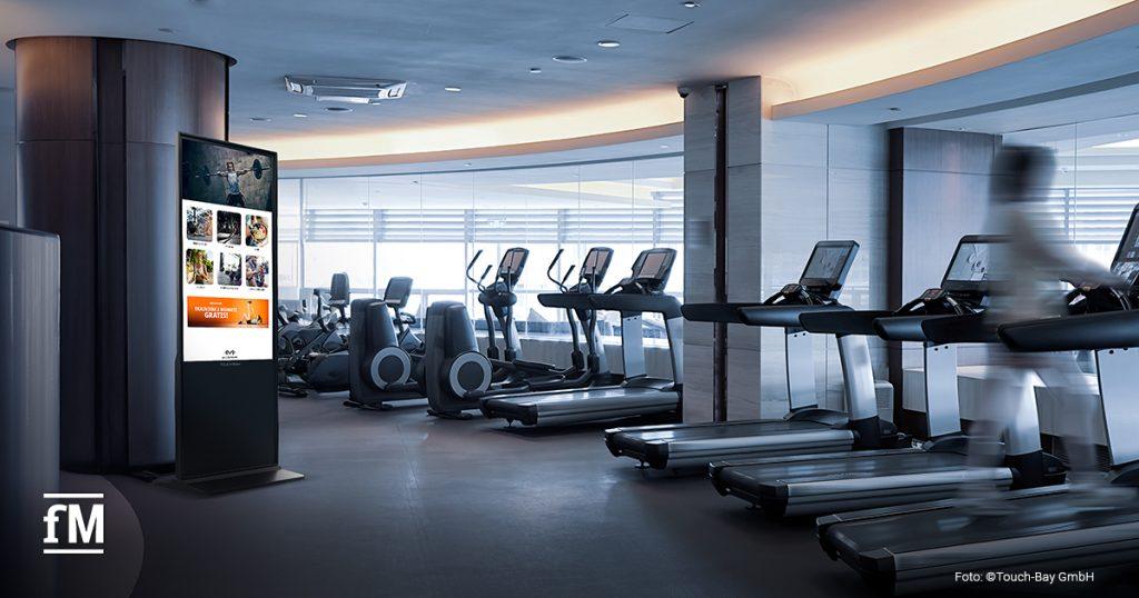 Hohes TOUCH:BAY Display mit Fitnessinhalten in einem großen Studio mit mehreren Laufbändern und moderner Beleuchtung.