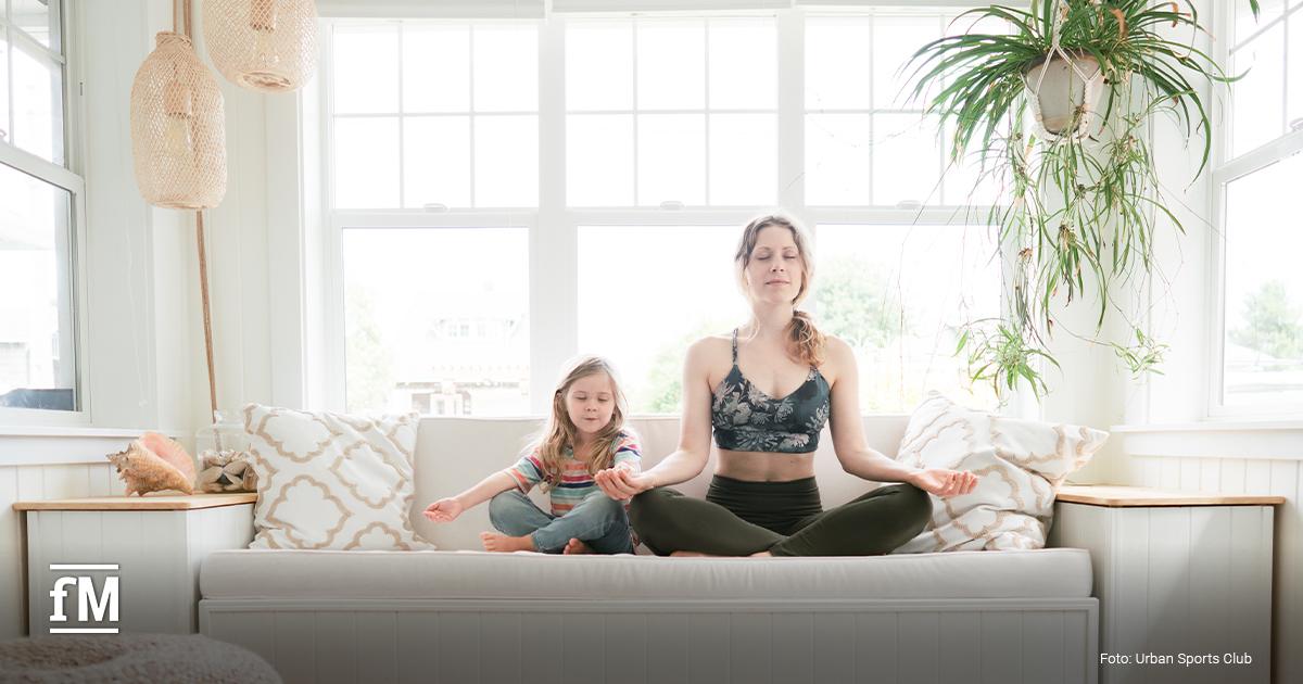 Frau und Kind sitzen im Schneidersitz meditierend auf einem Sofa in hellem Raum.