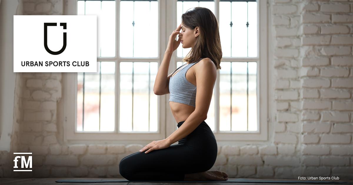 Frau im Sportoutfit sitzt auf Yogamatte und übt Atemtechnik in hellem Raum. Links oben ist das Logo von Urban Sports Club zu sehen.