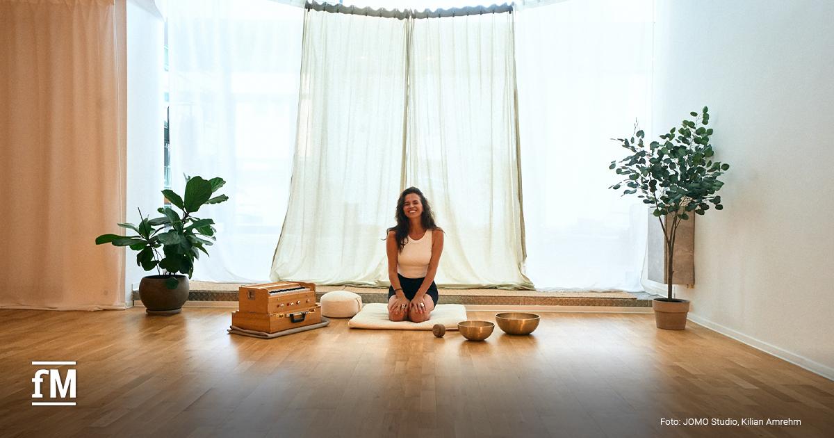 Frau sitzt im Yogastudio auf Matte, mit Musikinstrumenten und Pflanzen im Hintergrund.