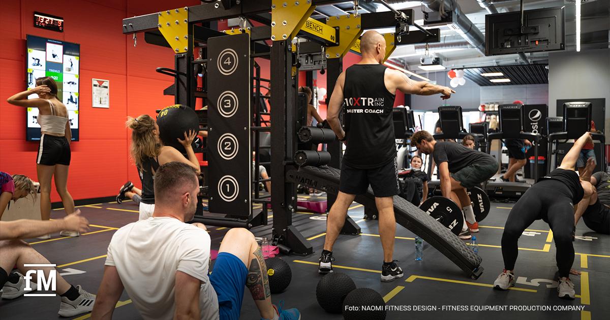 Coach steht mit dem Rücken zur Kamera in einem Fitnessstudio vor einer Trainingsgruppe und gibt Anweisungen, während mehrere Personen an funktionellen Stationen trainieren. Auf seinem schwarzen Muskelshirt ist die Aufschrift „10XTraining” zu lesen.