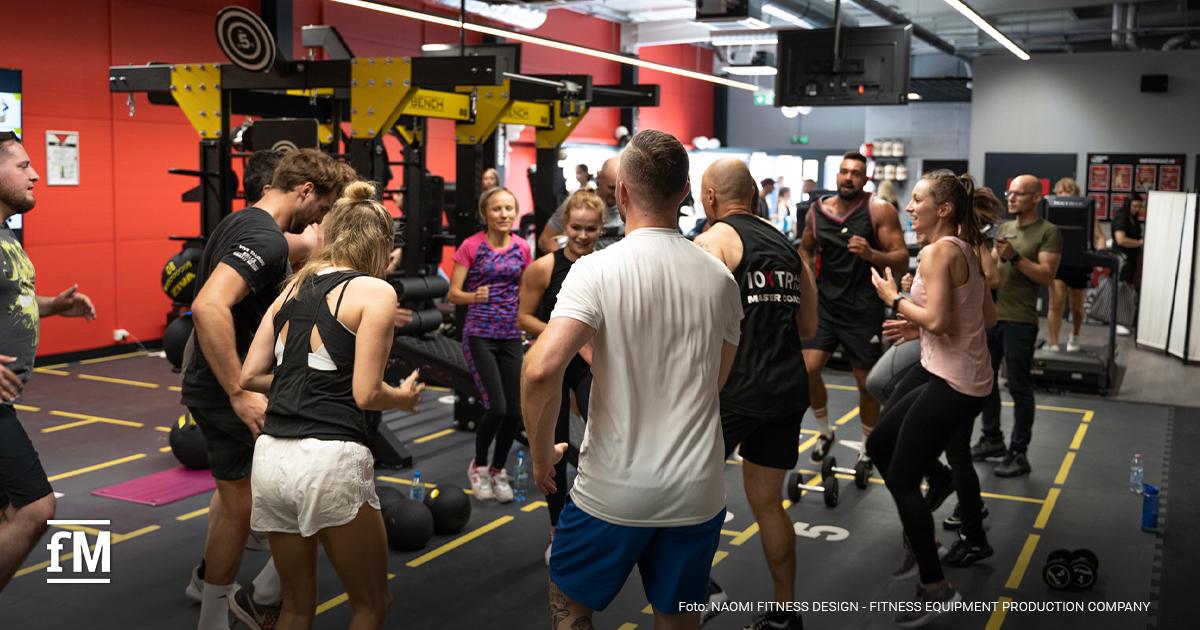 Trainingsgruppe bewegt sich gemeinsam in einem Fitnessstudio, Personen tragen Sportkleidung und nutzen eine offene Trainingsfläche.