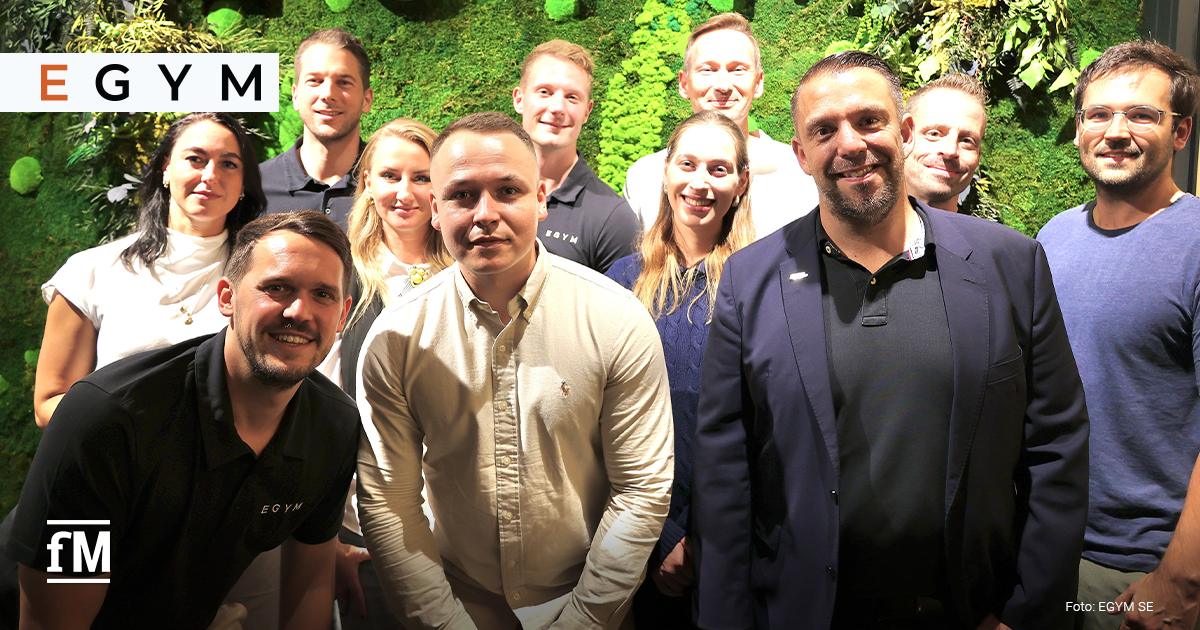 Eine Gruppe von Frauen und Männern steht vor einer begrünten Wand mit dem Schriftzug 'EGYM' und blickt in die Kamera.