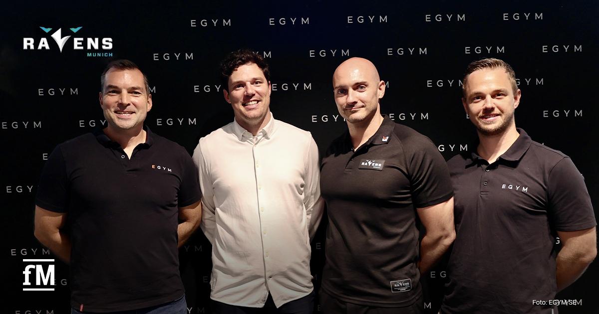 Vier Männer stehen nebeneinander vor einer schwarzen Wand mit EGYM-Logos, zwei tragen EGYM-Shirts, einer Munich Ravens-Kleidung