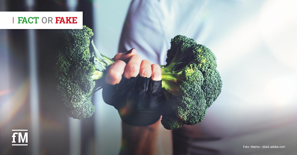 Eine Hand mit schwarzem Trainingshandschuh hält zwei Brokkoliköpfe wie eine Hantel vor hellem, unscharfem Hintergrund.