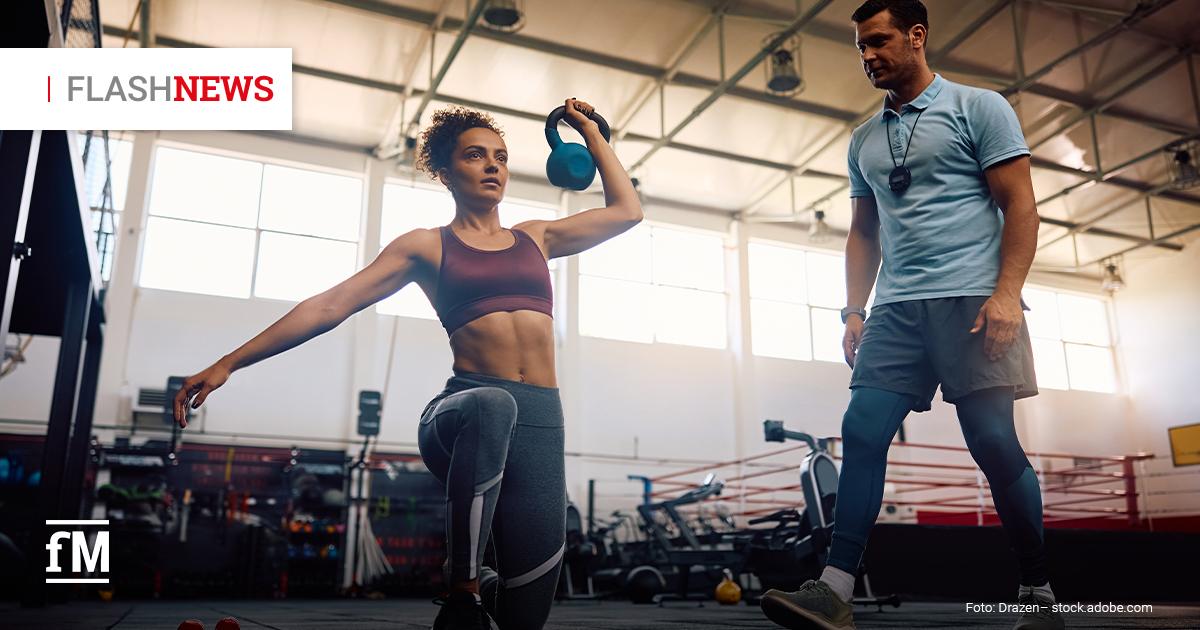 Eine Frau führt mit einer Kettlebell eine Ausfallschrittübung aus, während ein Trainer sie in einem Fitnessstudio beobachtet und anleitet.