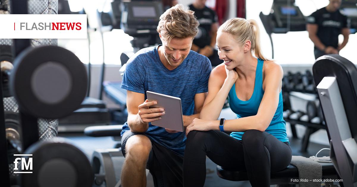 Ein sportlich gekleideter Mann und eine Frau sitzen im Fitnessstudio nebeneinander auf einer Hantelbank und schauen gemeinsam lächelnd auf ein Tablet. Im Hintergrund sind Fitnessgeräte und weitere trainierende Personen zu sehen.