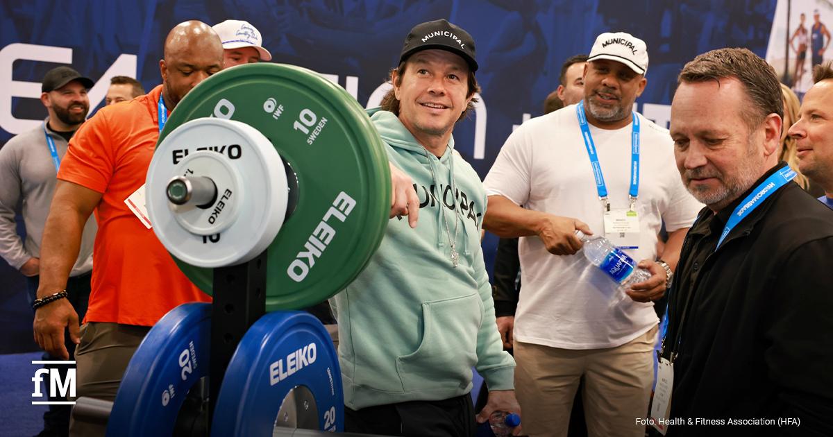 Messehalle im San Diego Convention Center mit mehreren Personen, Gewichten und Ausstellungsflächen im Vordergrund. Das Bild zeigt den Schauspieler Mark Wahlberg bei der HFA Show 2025 an einem Stand des schwedischen Gewichthebe-Ausrüstungsherstellers Eleiko. Er trägt einen mintgrünen Kapuzenpullover und eine schwarze Mütze mit der Aufschrift 'Municipal'.