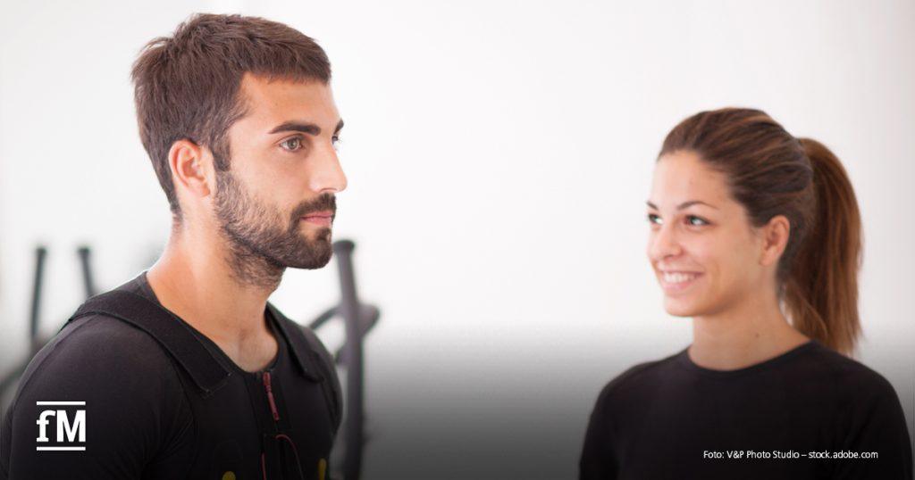 Zwei Personen in Trainingskleidung stehen sich gegenüber, eine Person trägt einen EMS-Anzug, beide befinden sich in einem Studio.