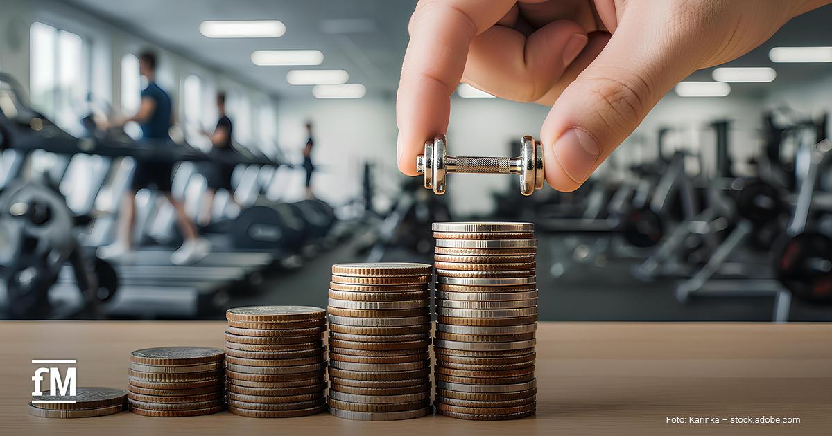 Die Hand hält eine kleine Hantel über gestapelte Münzen auf einem Tisch. Im Hintergrund sind unscharf Fitnessgeräte und trainierende Personen zu erkennen.