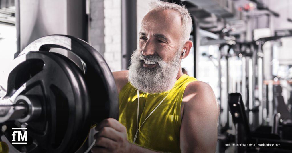 In einem Fitnessstudio hebt ein grauhaariger Mann mit weißem Bart eine Hantelscheibe an einer Kraftmaschine; Trainingsgeräte und helle Wände im Hintergrund