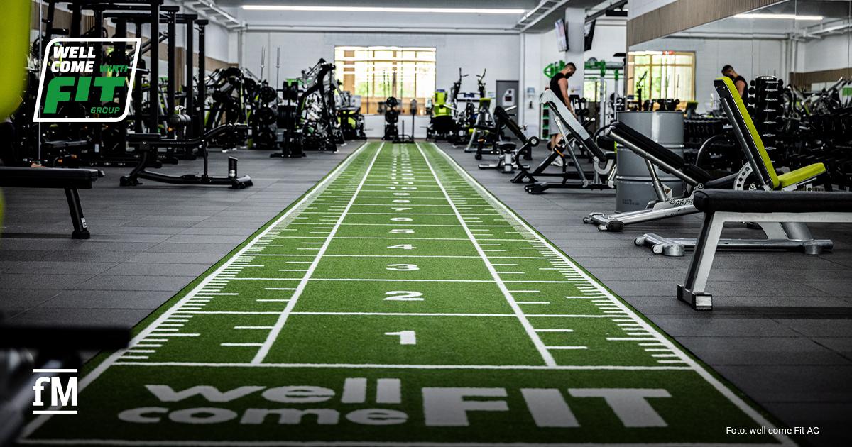 WELL COME FIT Übernahme Trainingsbereich in einem Fitnessstudio mit grünem Kunstrasen, Kraftgeräten, Hantelbänken und mehreren Trainingsmaschinen in einer großen Halle.
