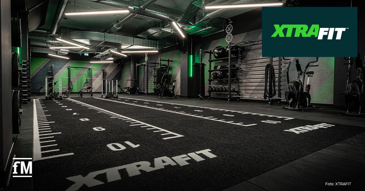 Großer Fitness-Trainingsraum mit Kunstrasenbahn, Markierungen und dem Schriftzug XTRAFIT, Kraftstationen, Gewichten, Seilen und moderner Beleuchtung.