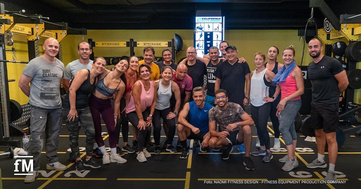 Trainingsgruppe aus Frauen und Männern in einem Fitnessclub, gemeinsam posierend auf einer markierten Trainingsfläche mit Geräten im Hintergrund.