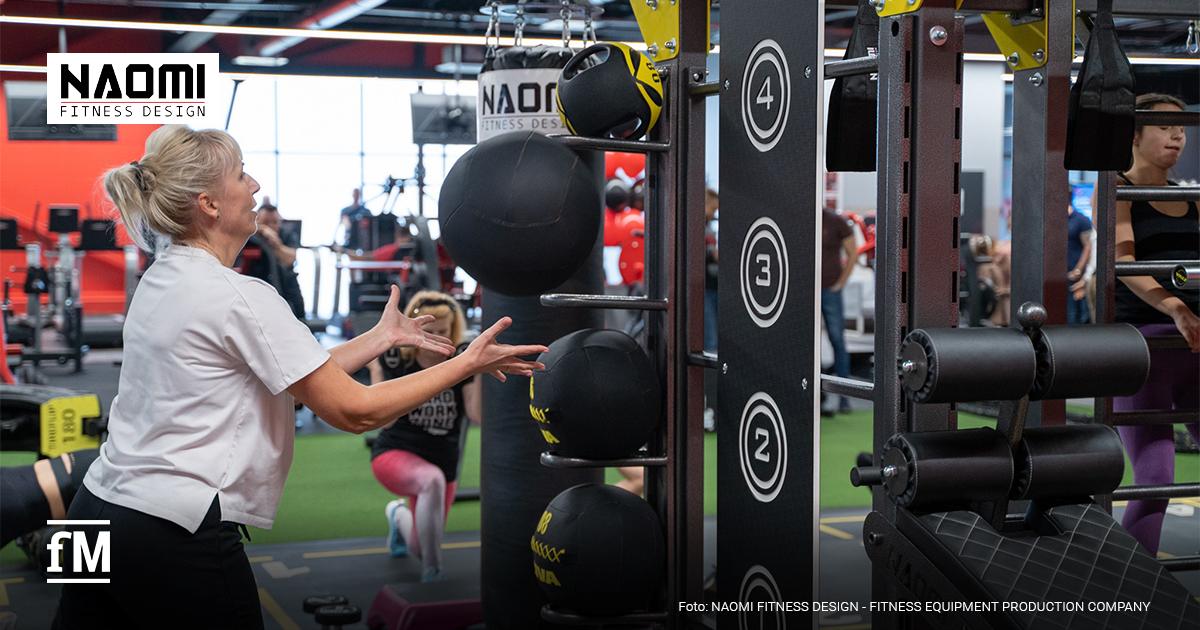 10XTraining Mitgliederbindung Fitnessclubs Frau wirft einen Medizinball in einem Fitnessstudio, weitere Trainierende und funktionelle Trainingsgeräte sind im Hintergrund zu sehen.