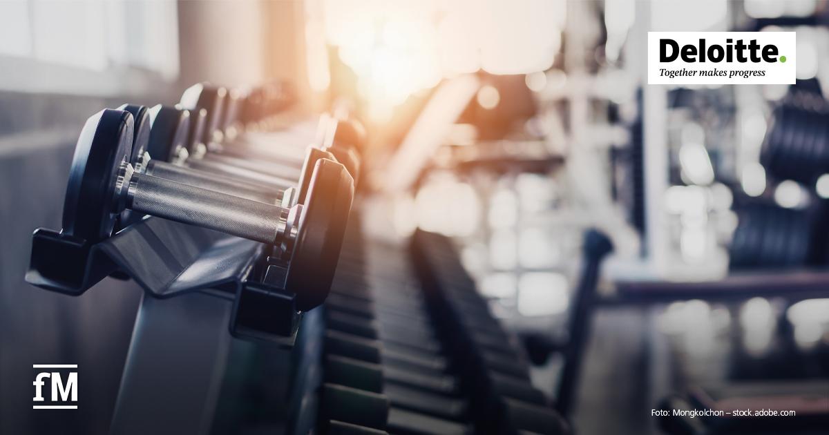 Im Vordergrund ist eine Hantelreihe auf schwarzem Rack abgebildet, Metallgriffe sind horizontal ausgerichtet; im Hintergrund ein unscharfer Fitnessraum mit Geräten und Fenstern.