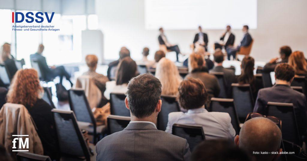 Konferenzraum aus hinterer Zuschauerperspektive; im Vordergrund mehrere sitzende Zuhörer auf Reihenstühlen mit Blick nach vorn, im Hintergrund Podium mit vier diskutierenden Personen vor Projektionsfläche