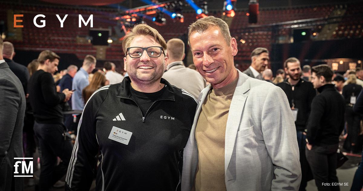 EGYM Kongress 2026 Zwei Männer, EGYM CEO Philipp Roesch-Schlanderer und TV Moderator Markus Othmer, in Businesskleidung nebeneinander vor der Bühne des EGYM-Kongresses lächeln in die Kamera. Im Hintergrund ist eine große Veranstaltungshalle mit weiteren Gästen zu sehen.