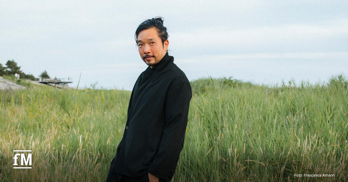 Mann in dunkler Kleidung stehend in hohem Gras in einer offenen Landschaft unter freiem Himmel