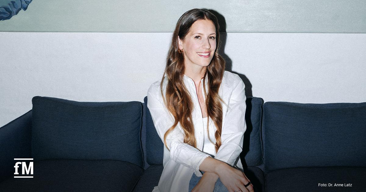 Sitzende Frau in heller Bluse und Jeans auf einem Sofa vor einer abstrakten Wandgestaltung in einem Innenraum