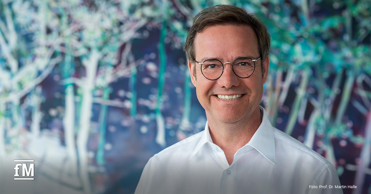 Mann mit Brille und weißem Hemd vor einem farbigen, unscharfen Hintergrund in einem Innenraum