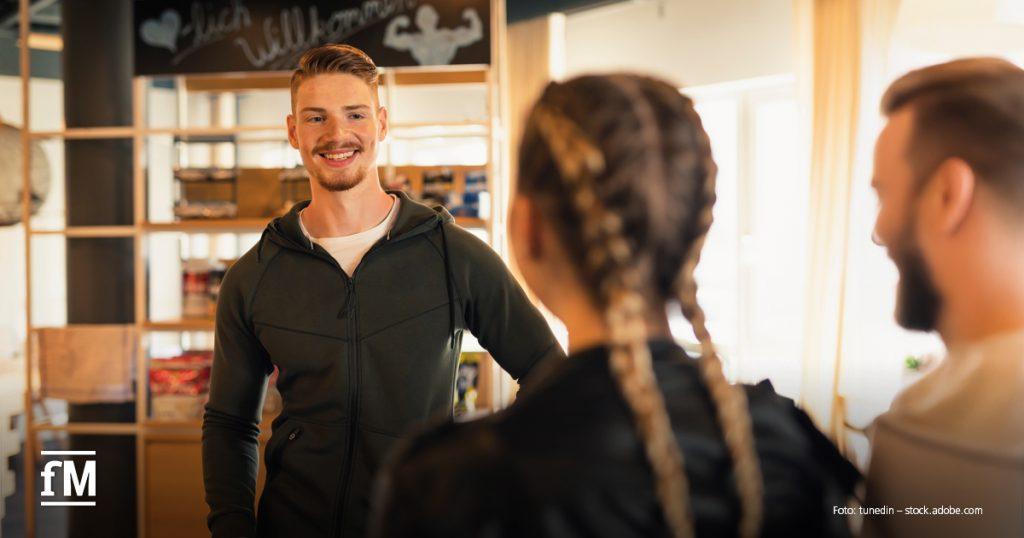 Mann im dunklen Trainingsoberteil steht im Fitnessstudio und spricht mit zwei Personen im Vordergrund; Regale und Schriftzug „Willkommen“ im Hintergrund.