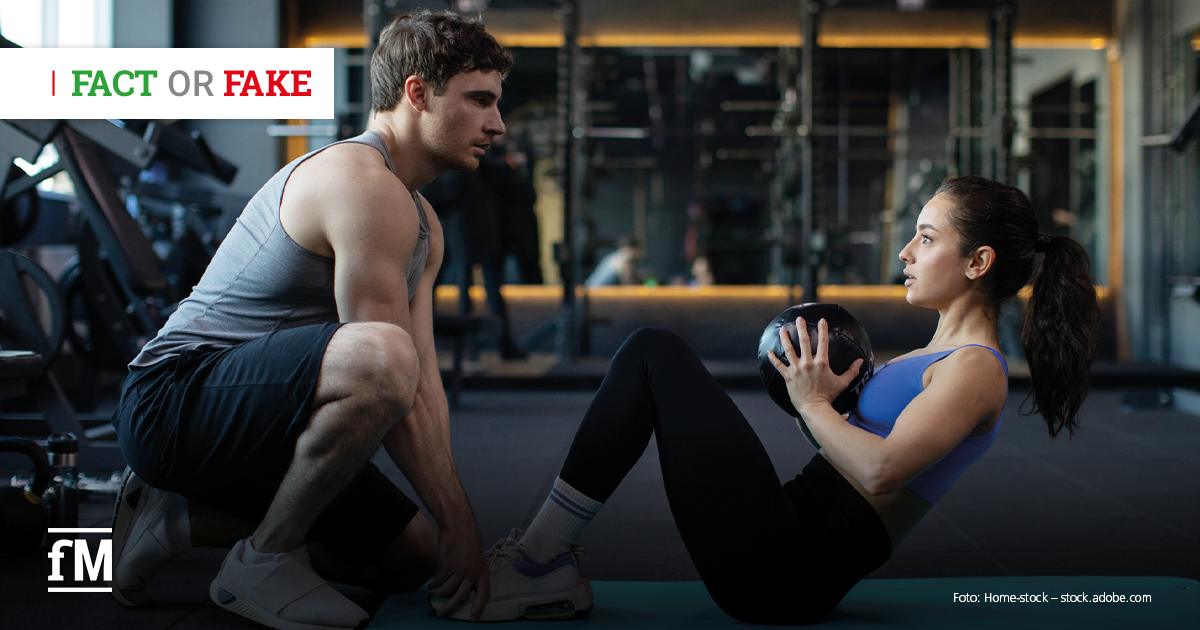 Im Vordergrund kniet links ein Trainer auf dem Boden und blickt nach rechts zu einer Frau, die auf einer Trainingsmatte sitzt und einen Medizinball vor der Brust hält; im Hintergrund sind Fitnessgeräte und Spiegel im Studio zu sehen.