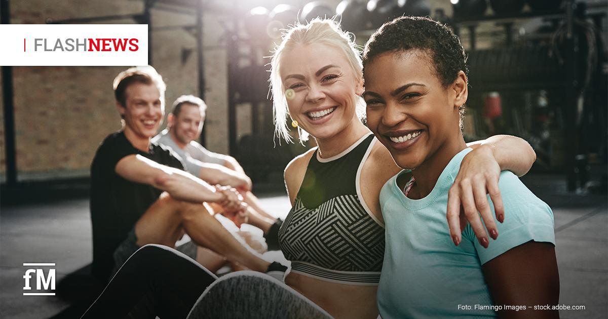 Fitness Flash News #2/2026 KW 9 Zwei Frauen sitzen lächelnd im Fitnessstudio, eine legt den Arm um die andere, im Hintergrund zwei weitere Personen auf Trainingsmatten, Schriftzug „FLASH NEWS“ oben links.
