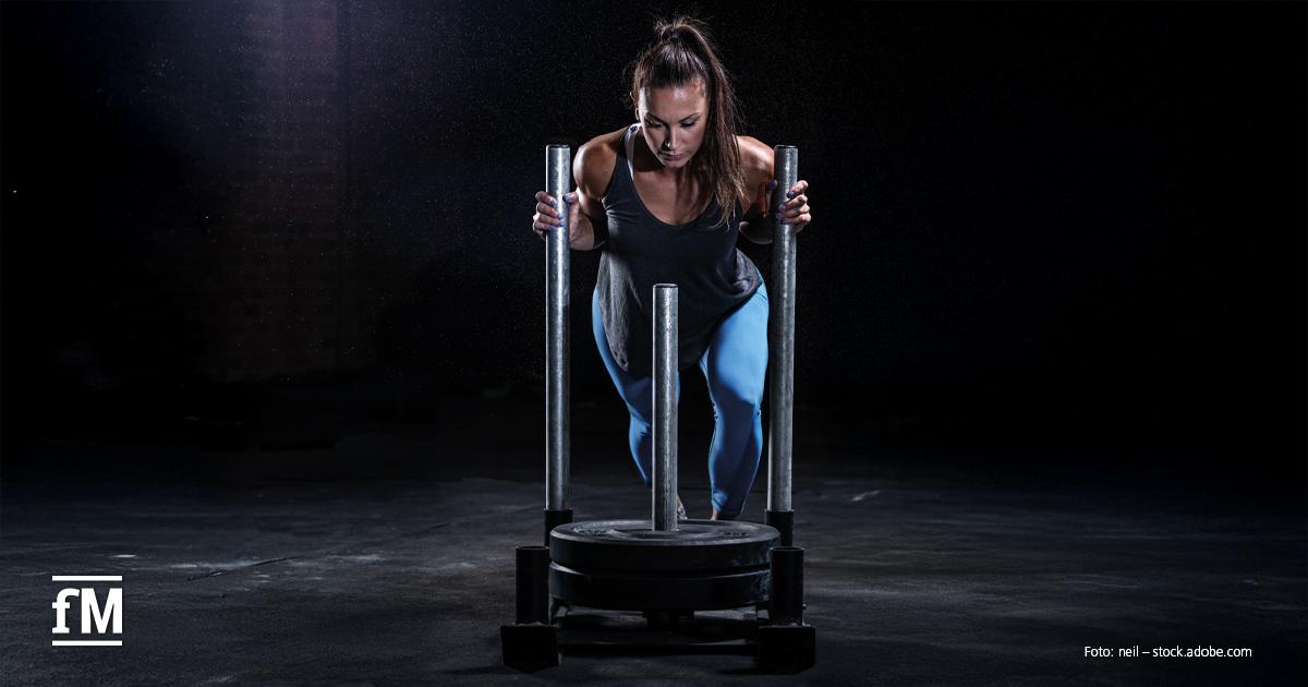 HYROX-Trend in Fitnessstudios Zentrale Perspektive auf eine Athletin in Vorwärtsbewegung, die einen Gewichtsschlitten auf einer dunklen Trainingsfläche schiebt