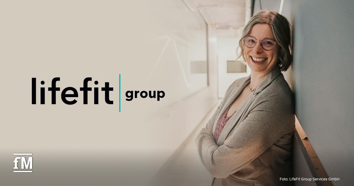 LifeFit Group neue HR-Direktorin Miriam Stein Flur eines modernen Bürogebäudes. Eine Frau mit blonden Haaren und Brille (Miriam Stein) lehnt mit verschränkten Armen an einer Wand neben einer Glasfront mit Lichtinstallation.