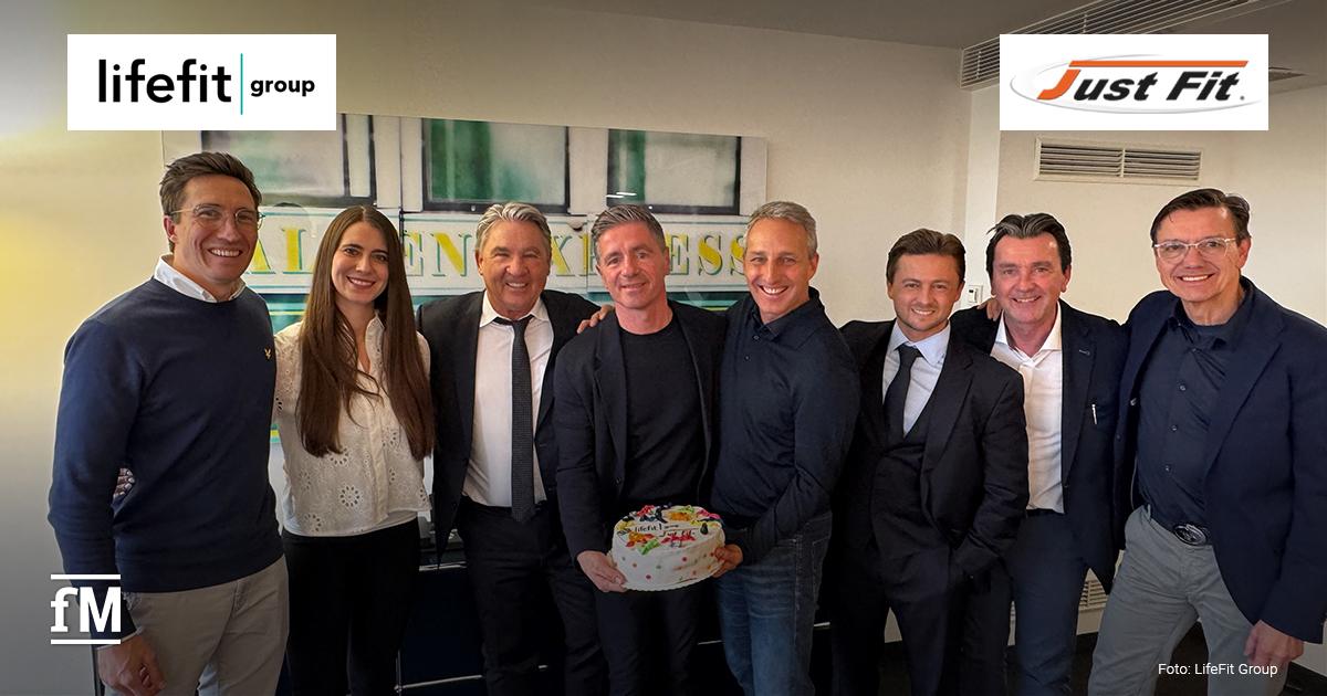 LifeFit Group Übernahme Just Fit Gruppenaufnahme in einem Innenraum in Köln mit mehreren Personen aus dem Management von Just Fit und der LifeFit Group, Logos beider Unternehmen im oberen Bildbereich.