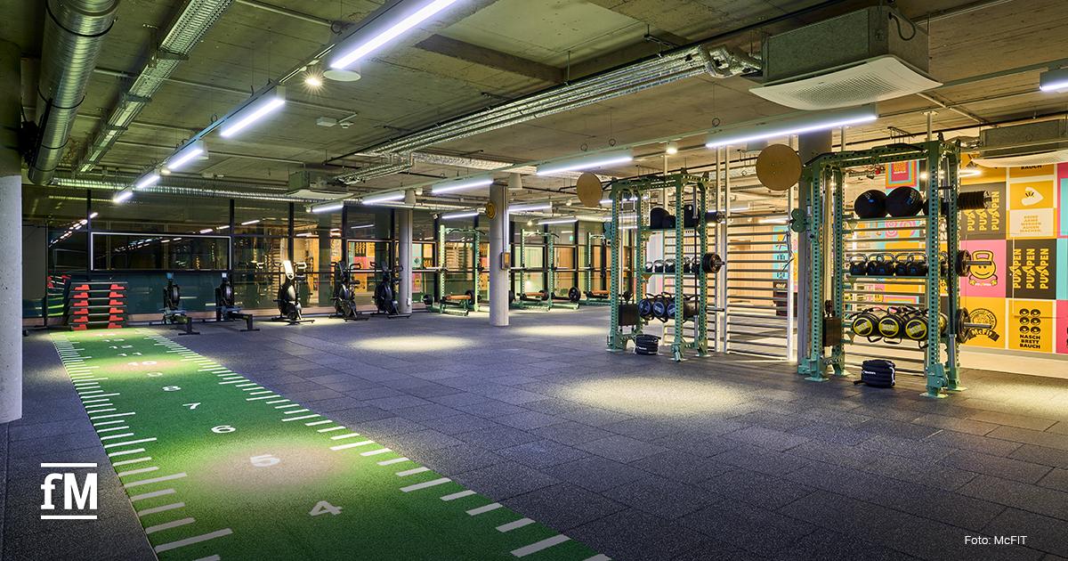 Trainingsfläche mit grün markierter Sprintbahn, Kraftstationen, Rudergeräten, Hanteln und Betonstützen in einem Fitnessstudio.