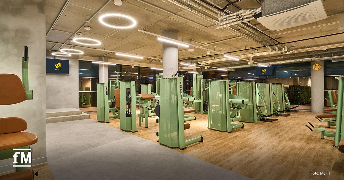 Trainingsraum mit grünen Kraftmaschinen auf Holz- und Betonboden, Betonstützen, Ringlichtern an der Decke und McFIT Bildschirmen.