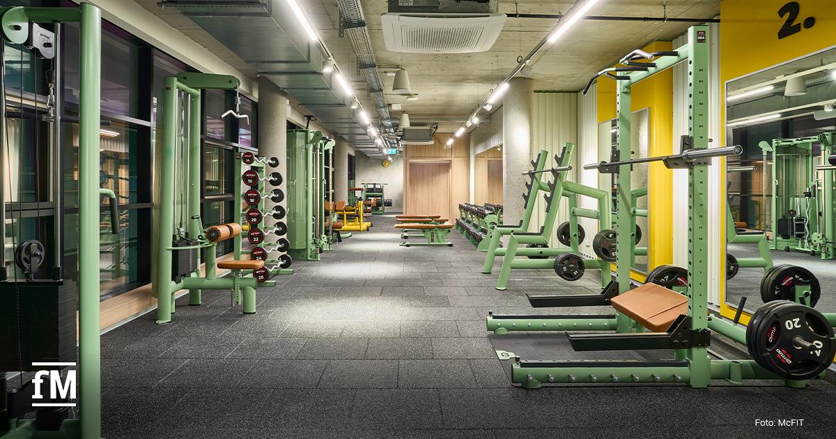 Fitnessstudio mit grünen Kraftstationen, Kurzhantelregal, Trainingsbänken, gelber Wand mit Zahl „2.“ und Fenstern links.