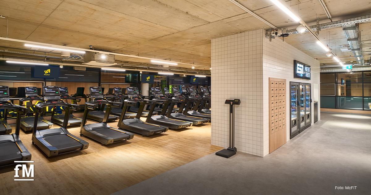 Reihe von Laufbändern auf Holzfußboden, rechts Spinde und Getränkeautomaten, Betondecke mit Beleuchtung und McFIT Bildschirme im Hintergrund.