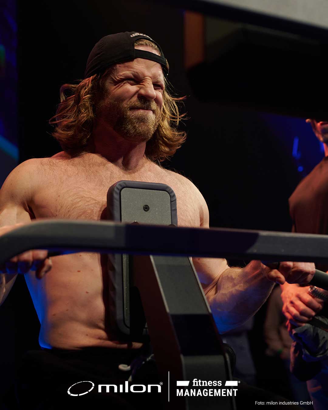 Oberkörperfreier Mann trainiert an einem Gerät auf Bühne, andere Personen stehen in der Nähe.