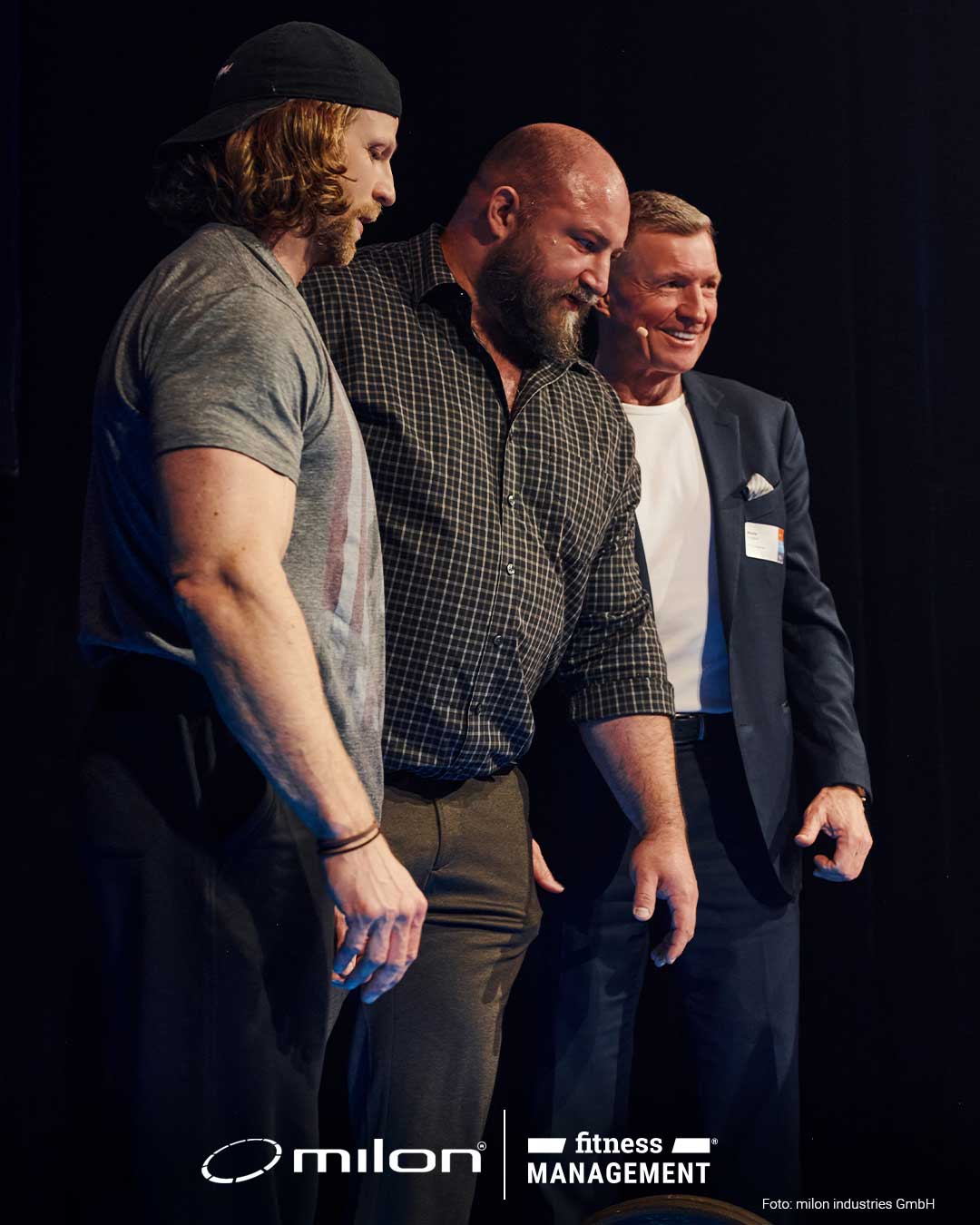 Drei Männer stehen nebeneinander auf einer Bühne und schauen nach unten, einer trägt ein Sakko.