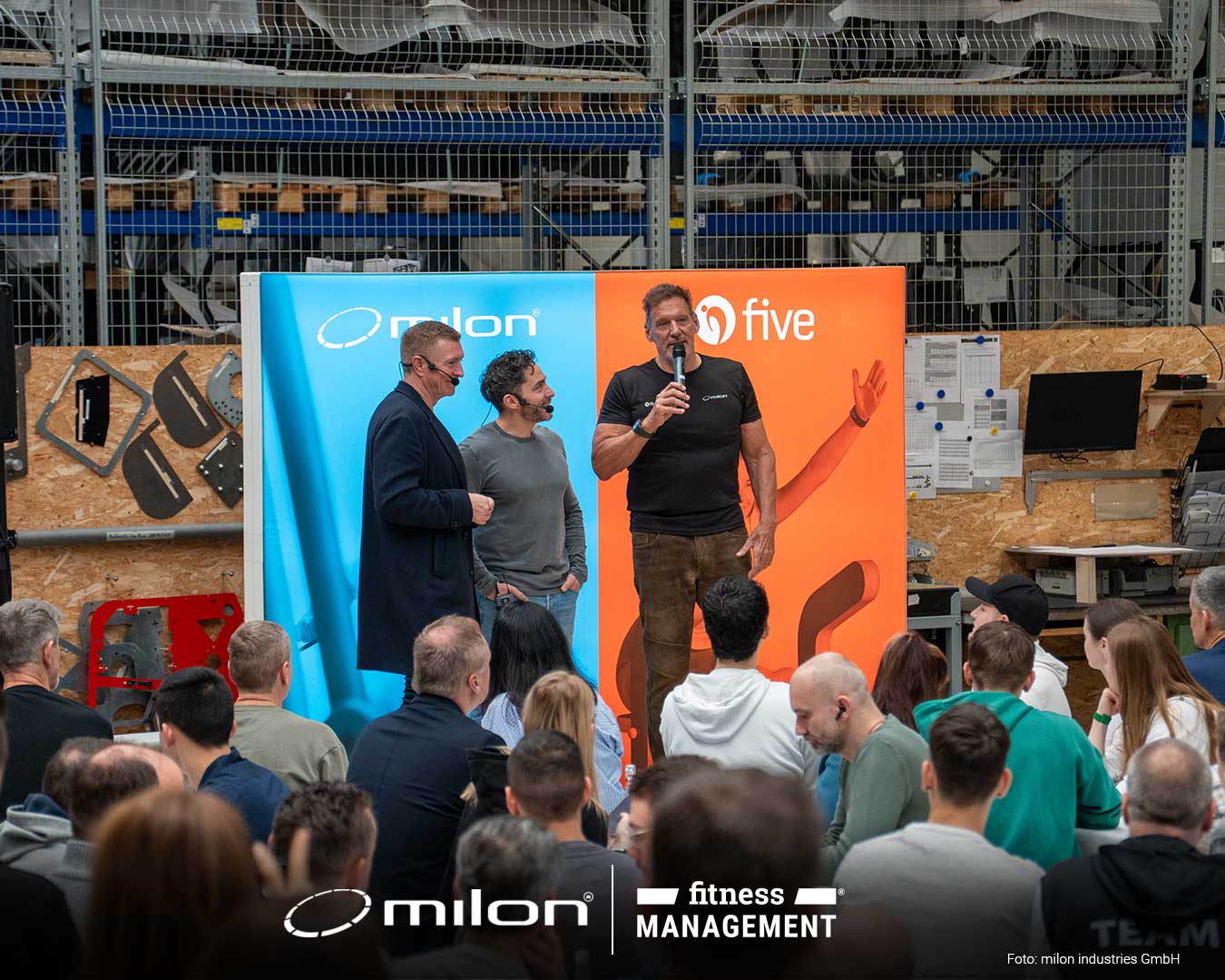 Drei Männer stehen vor einer milon und five Präsentationswand in einer Produktionshalle, Publikum sitzt davor.