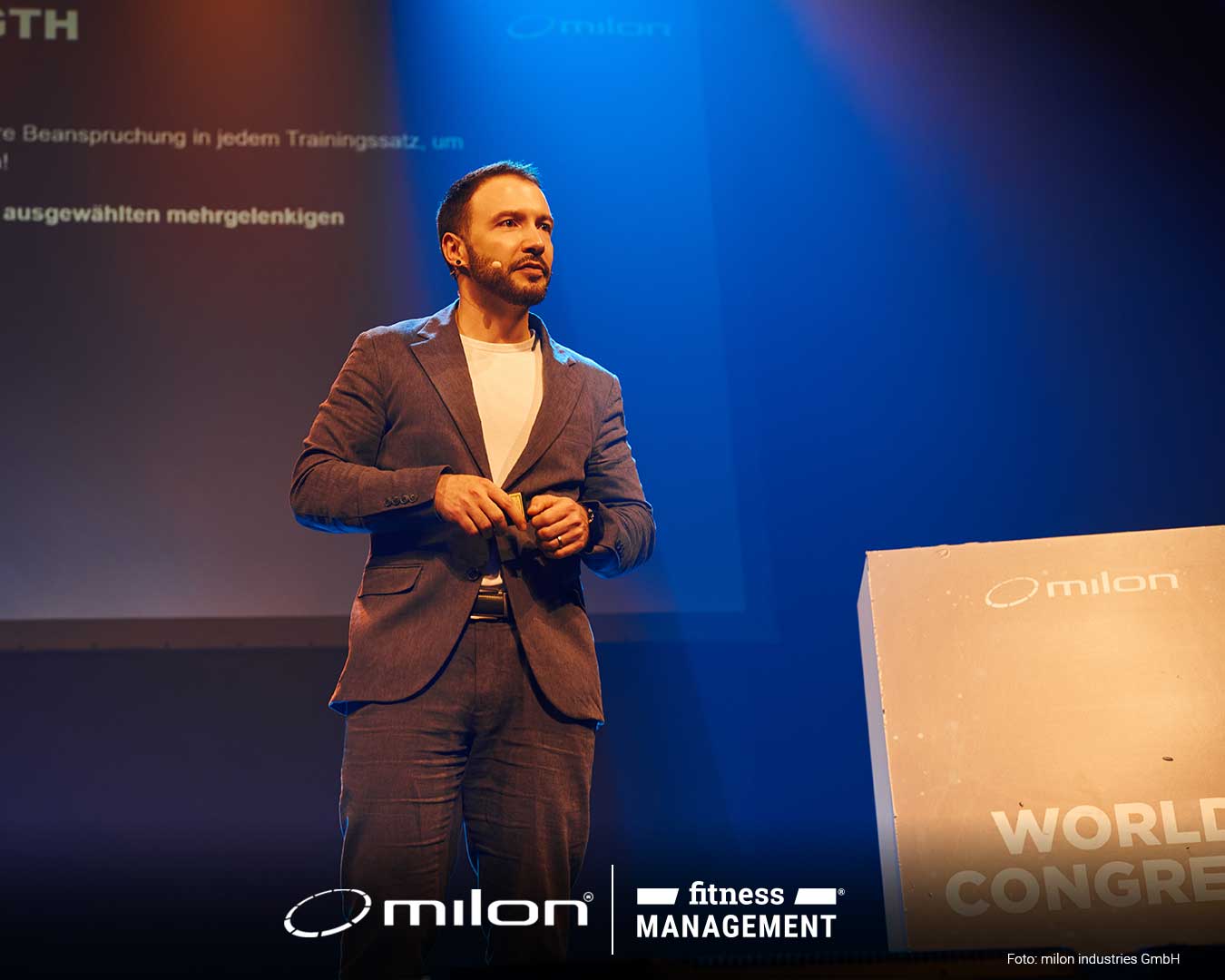 Mann im Anzug steht auf Bühne neben Rednerpult mit milon Logo, Präsentationsfolie im Hintergrund.
