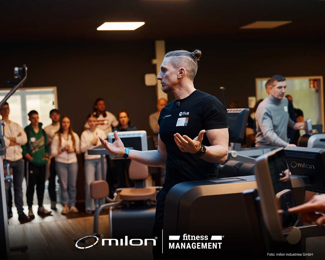 Mann im schwarzen Shirt spricht in Trainingsraum, mehrere Personen stehen im Hintergrund zwischen Geräten.