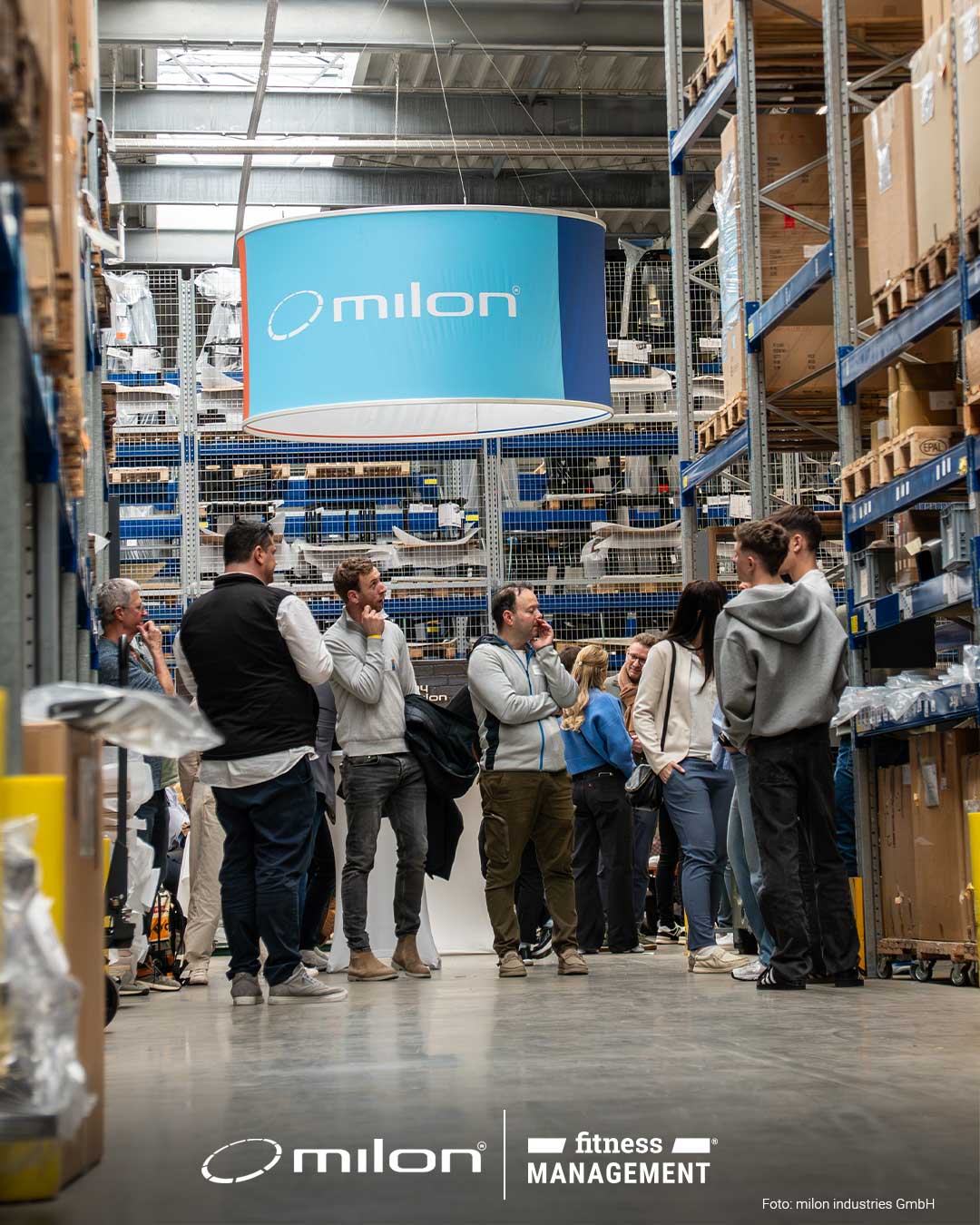 Gruppe von Personen steht in einer Lagerhalle zwischen Regalen mit Kartons, darüber ein rundes milon Schild.