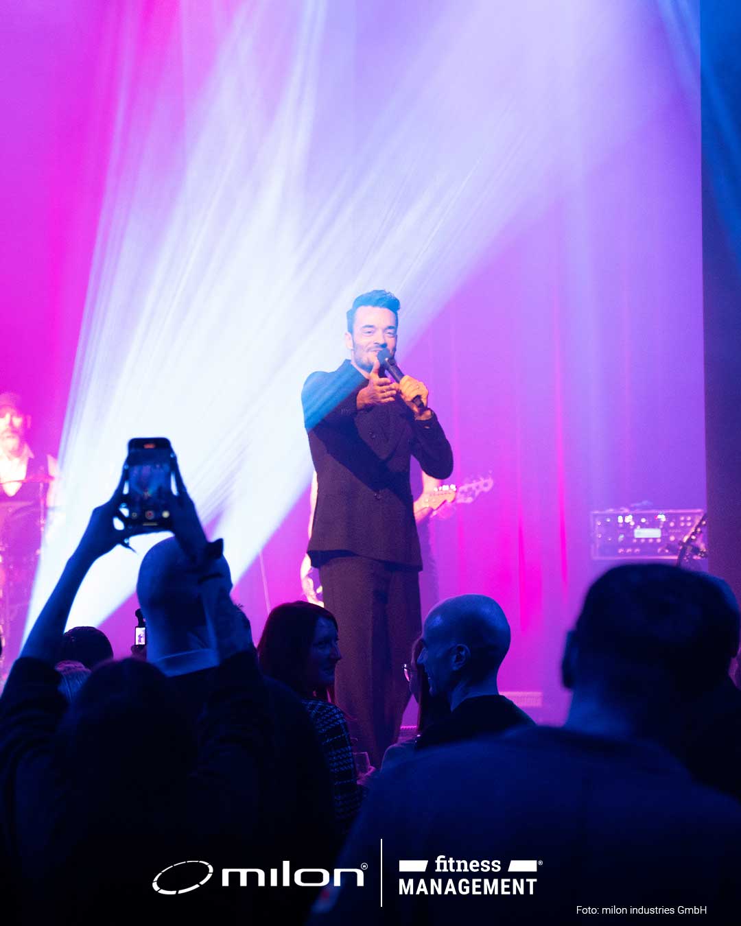 Sänger mit Mikrofon auf beleuchteter Bühne, Publikum im Vordergrund, farbige Scheinwerfer im Hintergrund.