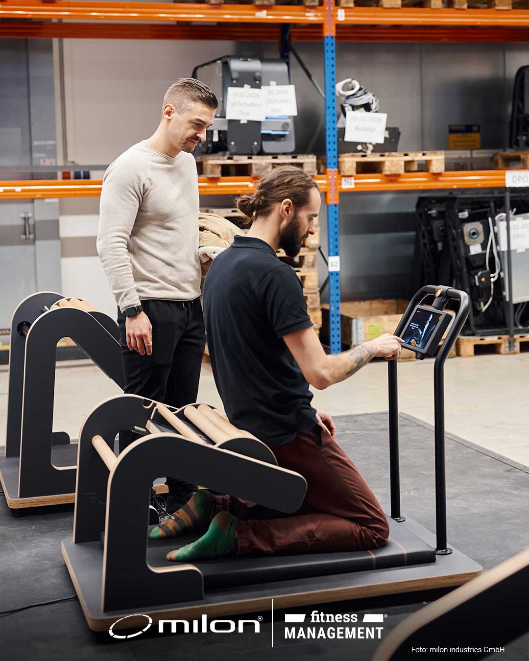 Zwei Männer testen ein Trainingsgerät in einer Halle mit Regalen und gelagerten Geräten im Hintergrund.