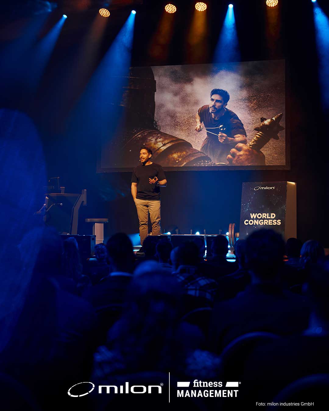 Mann steht auf Bühne vor großer Leinwand mit Bildprojektion, Publikum sitzt im dunklen Saal.