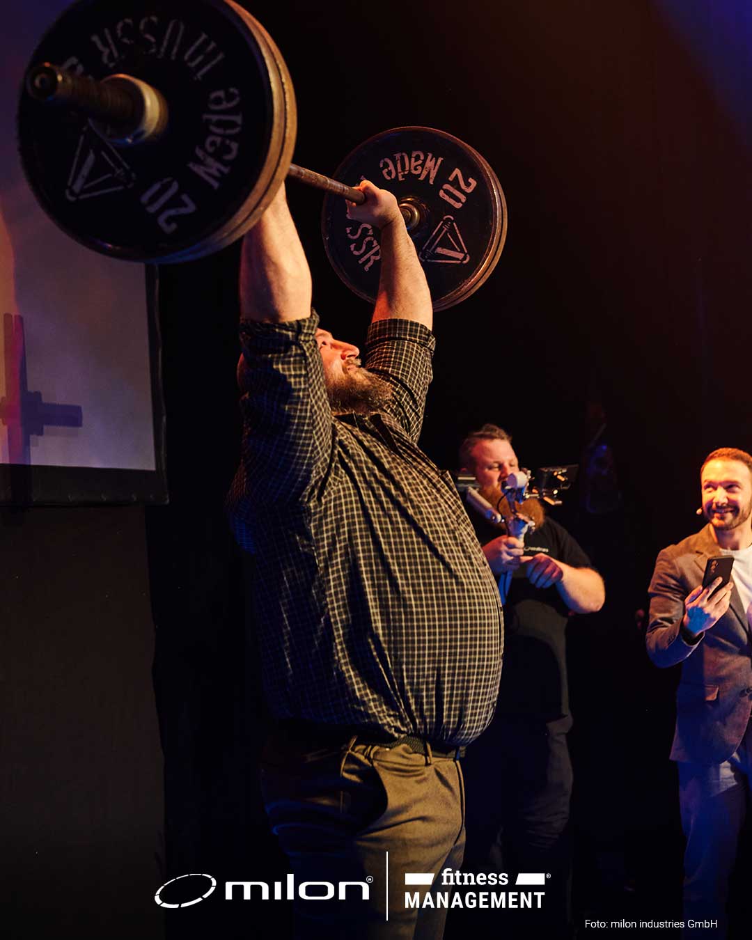 Mann hebt eine Langhantel über den Kopf auf einer Bühne, weitere Personen stehen daneben.