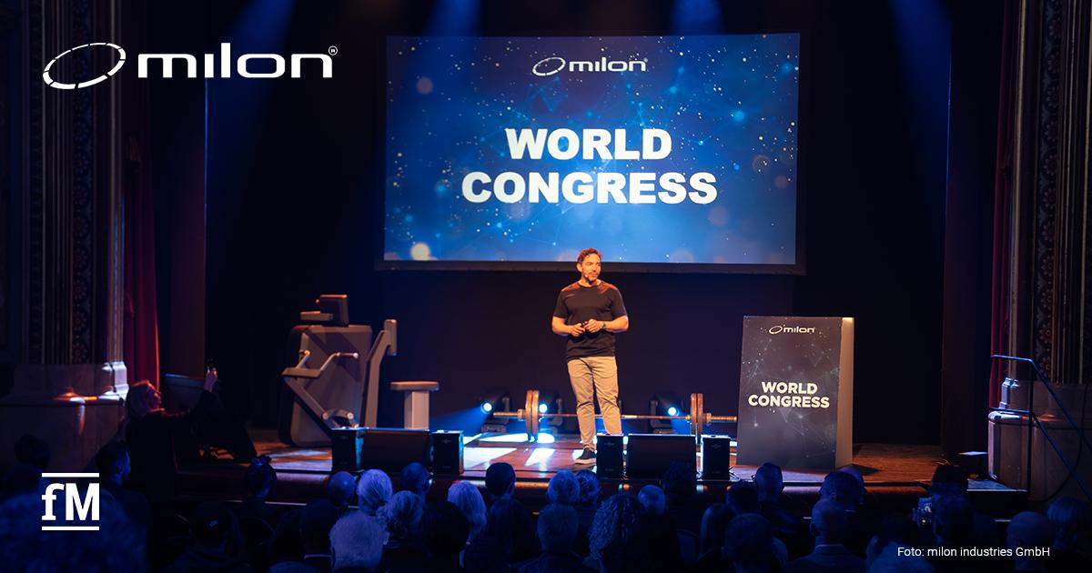 milon World Congress Mann steht auf Bühne vor großer Leinwand mit Schriftzug „WORLD CONGRESS“, Publikum sitzt im Saal.