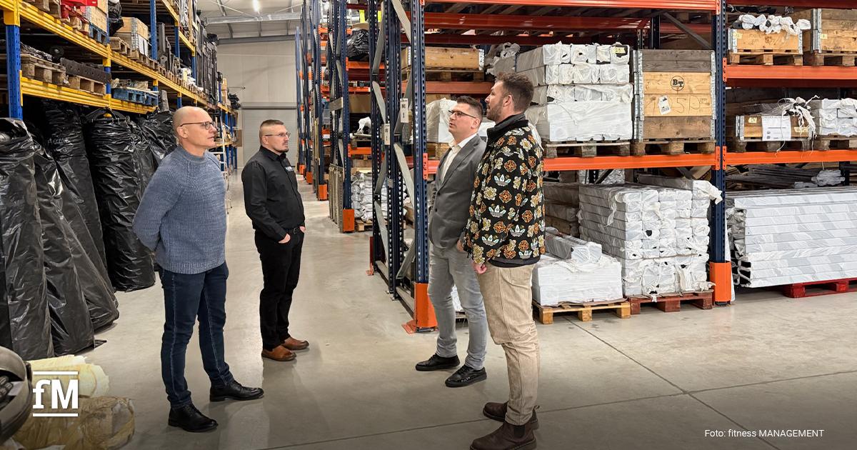 Große Lagerhalle mit hohen Metallregalen, verpackten Bauteilen auf Paletten und vier Personen im Gespräch im Mittelgang.