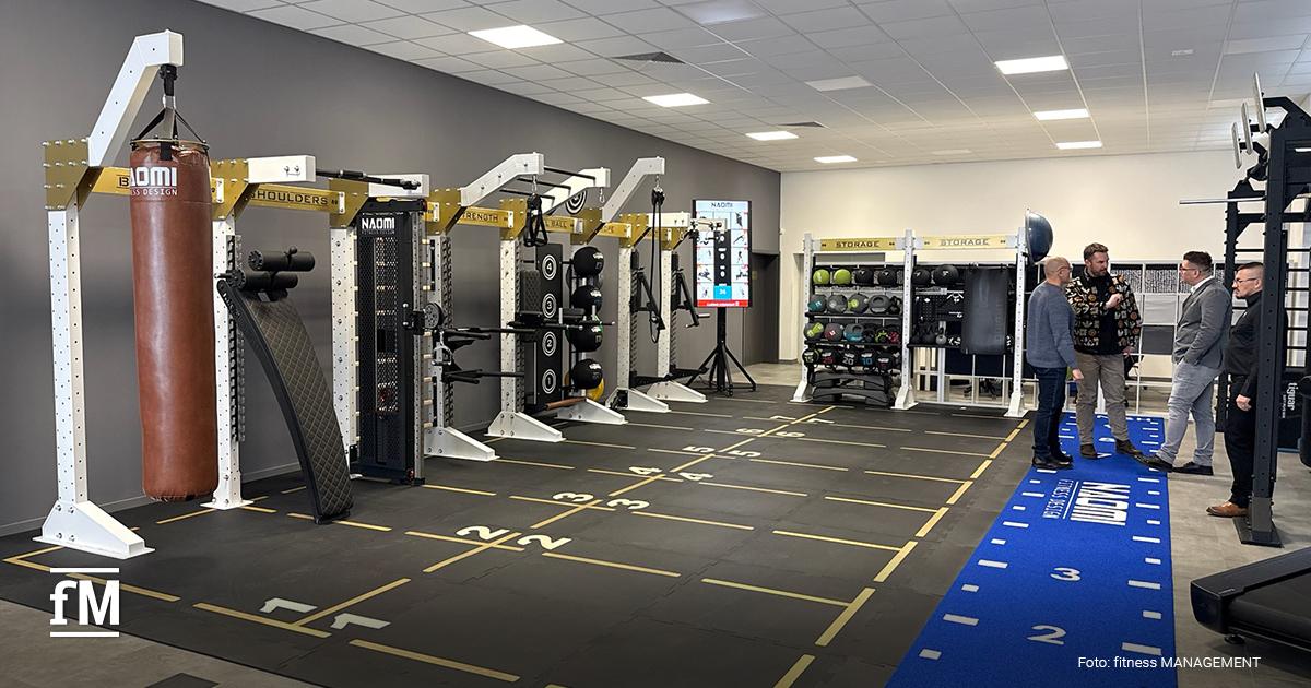 Trainingsraum mit weißem Functionalrig, Boxsack links, gelb markierte Bodenfelder und drei Personen im Gespräch rechts.