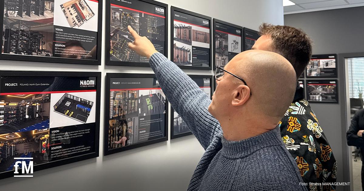 Wand mit gerahmten Projektplänen und Studioabbildungen, zwei Männer zeigen auf einzelne Layoutdarstellungen.