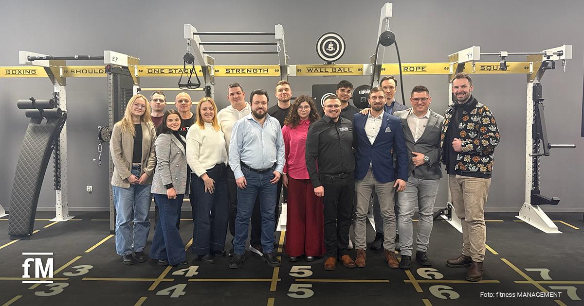 Mehr als zehn Personen stehen in einer Reihe vor einem Functionalrig auf markierten Bodenfeldern, graue Wand im Hintergrund.